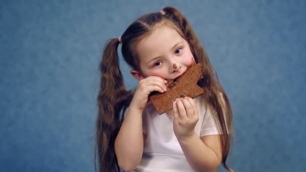 チョコレートを食べる少女の肖像画 ポニーテールが付いているおかしい子供は 汚れた顔でお菓子を食べます 青いスタジオの背景に汚れた子供 — ストック動画