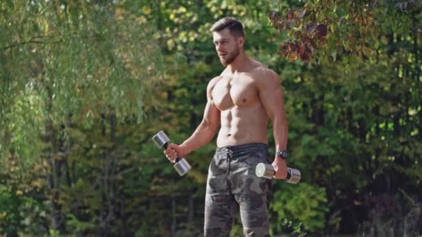 汚れたシャツのない男は 自然の中でトレーニングをしている 屋外ダンベルで運動する筋肉ボディービルダー ダンベルを持ち上げるハンサムアスリート — ストック動画