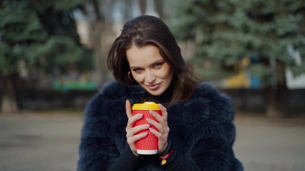 屋外の飲料と毛皮のコートの可愛い女の子 美しい女性の肖像画 ぼやけた公園の背景にあるプラスチック製のカップ — ストック動画