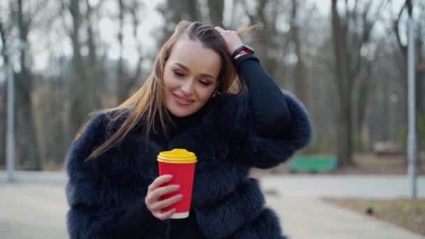 屋外にプラスチック製のカップが付いている美しい女性 公園を歩いている毛皮のコートの美女 公園内のブルネットの景色を眺める スローモーション — ストック動画