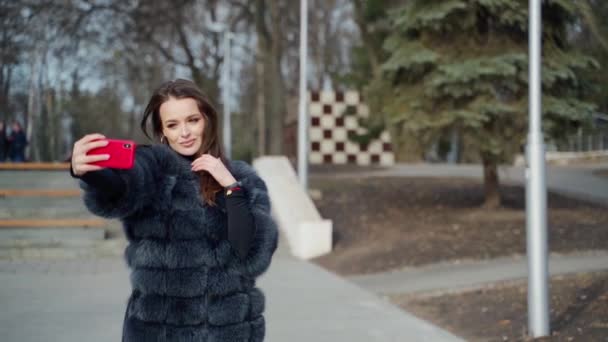 公園でセルフィーをしている美しい女の子の肖像画 高級毛皮コートの若い女性は晴れた冬の日に赤い電話で自分自身の写真を撮ります — ストック動画