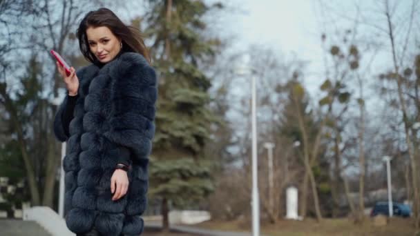 穿着毛皮大衣的奢华女人 漂亮的年轻模特拿着电话在公园里散步 在寒冷的季节看着相机 慢动作 — 图库视频影像