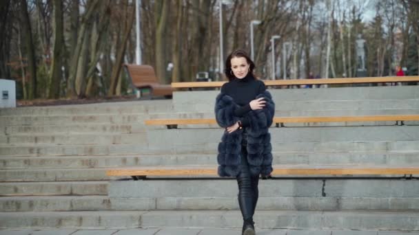 秋の公園でカメラにポーズする美しいモデル 黒い毛皮のコートを着た若い女性が歩き カメラを見ている スローモーション — ストック動画