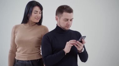 Genç mutlu çift stüdyoda telefona bakıyor. Siyah kazaklı adam elinde bir cep telefonu ve arkasında gülümseyen bir kadın tutuyor. Yavaş çekim.