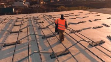 İki işçi çatıya metal temeli kuruyor. Koruyucu üniformalı teknisyenler güneş panelleri için yer hazırlıyorlar. Ağır çekim. Kamera hareket ediyor.