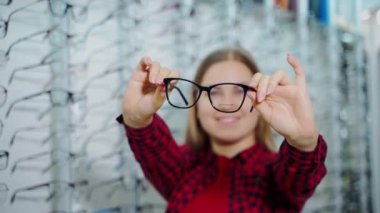 Kırmızı gömlekli güzel kız yeni gözlük seçiyor. Optik dükkanında elinde gözlüklerle gülümseyen çekici genç bir kadın..