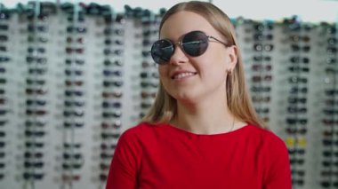 Güzel bir kadın şık güneş gözlüğü deniyor. Modern siyah güneş gözlüklü uzun saçlı kız optik mağazada kamera önünde poz veriyor..