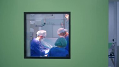 Bir grup cerrah ameliyat yapıyor. Ameliyathanede mavi üniformalı doktorlar var. Pencereden bak.