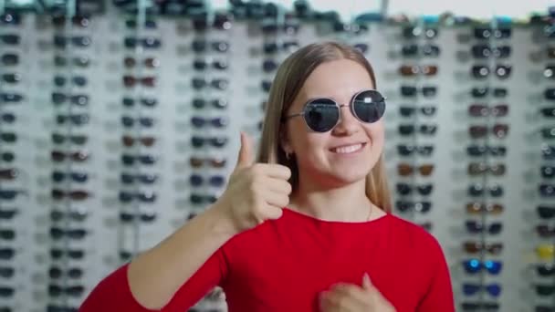 店内のファッショナブルなサングラスの可愛い女の子 光学のサングラスの背景に手でOkサインを示す素敵な少女の肖像画 — ストック動画