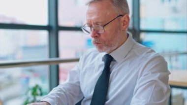 Dizüstü bilgisayarda çalışan düşünceli bir kıdemli adam. Olgun, gri saçlı bir iş adamının portresi. Pencerenin yanında oturuyor ve ofiste çalışıyor..