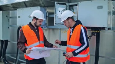 Mühendisler ileriki çalışmaları bir kağıt planıyla tartışıyor. Güvenlik kasklı erkek işçiler, arka plandaki elektrikli ekipman şemasına bakıyorlar..