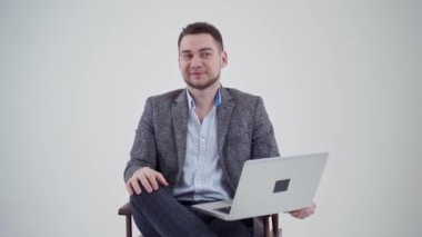 Bilgisayarı olan yakışıklı bir iş adamının portresi. Beyaz arka plandaki sandalyede oturan gri kostümlü gülümseyen genç adam. Yavaş çekim.