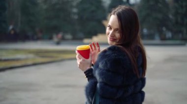 Açık havada kürk giymiş gülümseyen bir kızın portresi. Çekici görünümlü lüks bir kadın elinde bir fincan kahveyle parkta kameraya bakıyor..
