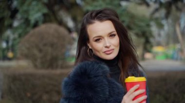 Lüks bir genç kadının yüzü. Çekici bir mankenin portresi, uzun siyah saçlı, kalın ceketli, elinde plastik bir fincan kahve olan ve parktaki kameraya bakan..
