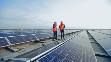 Gökyüzünün altındaki bir çatıda modern güneş çiftliği. Üniformalı ve koruyucu kasklı işçiler güneş panelleri kurulumunu tartıştılar.