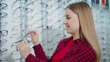 Optik dükkanında güzel bir sarışının yan görüntüsü. Genç bir kadın gözlüklü yeni moda gözlüklere bakıyor. Göz giysisi seçimi.