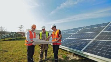 Güneşli bir günde güneşli bataryanın yanında teknisyenler. Koruyucu miğferli üç işçi güneş çiftliği planını inceliyor..