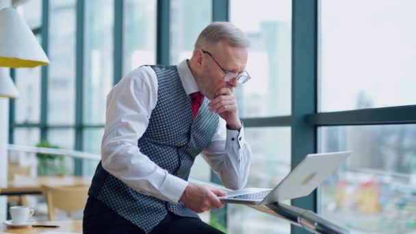メガネをかけた真面目なビジネスマンがラップトップを見つめている オフィスウィンドウの近くのラップトップで作業するエレガントなスーツの成熟したディレクター — ストック動画