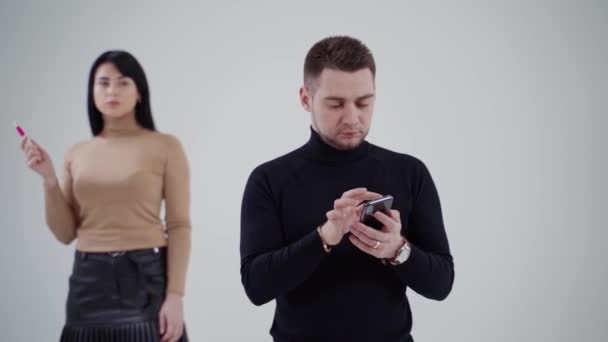 黒いセーターの若者は携帯電話をクリックします 男の後ろに立ってスタジオの背景で喫煙する魅力的な悲しい女性 — ストック動画