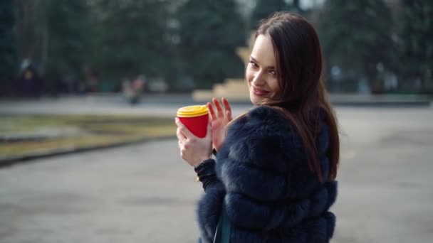 屋外の毛皮のコートの笑顔の女の子の肖像画 コーヒーの紙コップを持ち 公園でカメラを見ている魅力的な外観の豪華な女性 — ストック動画