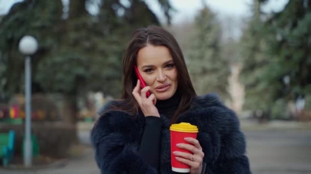 電話とホットコーヒーと紅茶のカップを持っているかわいい女性 高級毛皮コートの美しい女の子の肖像画 携帯電話を話し 公園でコーヒーを飲む — ストック動画