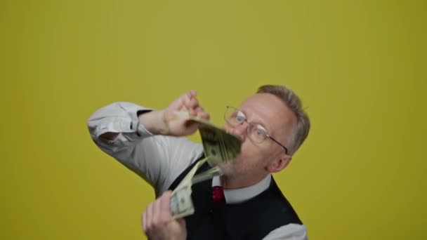 手元にお金を持ったシニアマン 幸せな金持ちは 黄色いスタジオの背景にドル紙幣を振っている 中年男性は大金を楽しんでいます — ストック動画