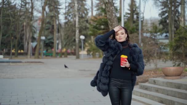 市立公園の高級毛皮のコートの美しい若い女の子 屋外でカメラを見ているコーヒーを飲む豪華な女性の素晴らしい景色 スローモーション — ストック動画