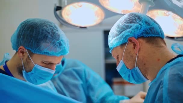 两名穿着外科制服的医生和女助手 在诊所的医疗灯下 外科医生在手术过程中戴着口罩 后续行动 — 图库视频影像