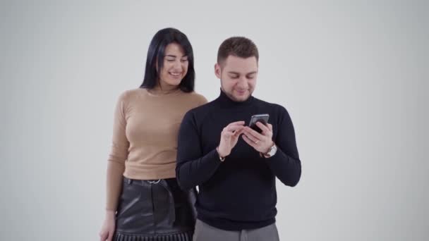 スタジオで笑顔の若いカップル ビジネスパートナーが一緒に立ち上がり 電話を眺めている 白い背景にハンサムな男と魅力的な女性 — ストック動画