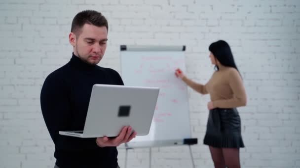 スタジオにいる若者と女性 女性が彼の後ろの白いボードに書いている間 ノートパソコンで作業するハンサムな男 — ストック動画