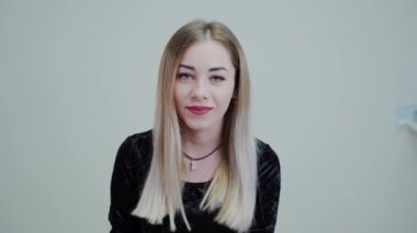 Beyaz arka planda oturan güzel sarışın kız gülümsüyor. Stüdyoda video.
