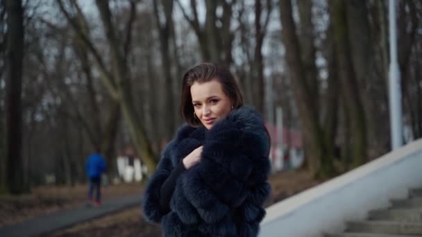 公園でポーズするスタイリッシュなダーク毛皮のコートを着用する若い美しいファッショナブルな女性 外を見ているモデル 女性ファッションコンセプトビデオ — ストック動画
