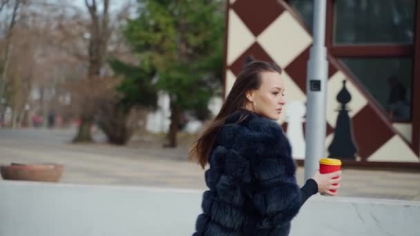 公園で歩くコーヒーと紙コップを保持する豪華な毛を身に着けているダークヘアの美しい官能的な少女 フラージュ ビデオ — ストック動画