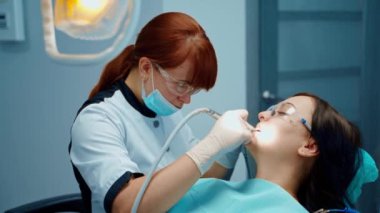 Genç bir kadın için dişçilik prosedürü. Maske ve gözlük uzmanı bir kadın modern klinikte diş teçhizatı ile hastanın dişlerini tedavi ediyor..