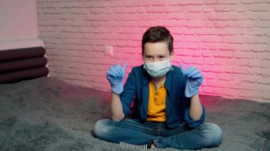 Çocuk izole edilmiş, koruyucu steril tıbbi maskeyle evde oturuyor. Yüzünde ve eldivenlerinde. Karantina, enfeksiyonu önleyin. Koronavirüsü durdurun. Evde kal.