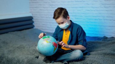 Kendini izole etmiş bir çocuk, Covid-19 'u öğrenen küreye bakıyor. Tıbbî maskeli bir çocuk. Gezegeni düşünüyor. Karantina, karantina ve sosyal uzaklık kavramı sırasında.