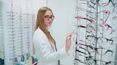 Optik mağazasında şık bir genç kadın. Beyaz ceketli güzel bayan yeni gözlük arıyor. Yuvarlak standın yanında gözlüklü bir alıcı..