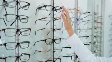 Gözlük, optik mağazadaki standın üzerinde. Modaya uygun gözlükleri seçen genç alıcı kadın. Göz giyimi konsepti.