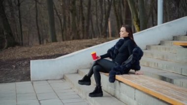 Açık havada bir fincan kahve içen seksi genç bir kadın. Soğuk mevsimde parkta bankta tek başına oturan ve çay içen güzel bir bayan. Yavaş çekim.