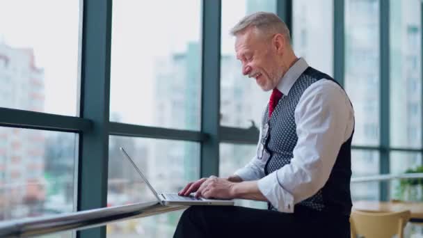 大きな窓の近くのラップトップにタイピングするハッピーシニアマン 自宅のコンピューターで働くフリーランサー リモート フリーランスは引退に取り組んでいます 高齢者のアクティブなモダンライフスタイル — ストック動画