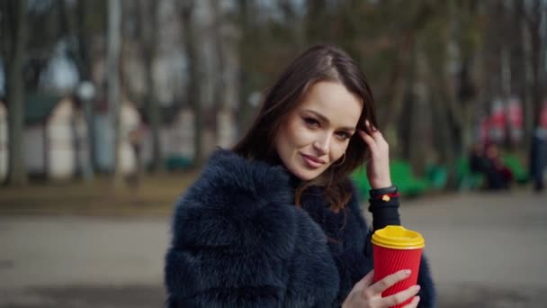 公園でコーヒーのプラスチックカップを持っている美しい女性 屋外でカメラにポーズする毛皮コートのブルネットガールの肖像画 スローモーション — ストック動画