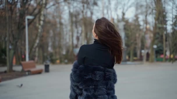 屋外でカメラを見ている長い髪のファッショナブルな女の子 毛皮コートの魅力的な若い女性の肖像画は 公園の背景にカメラにポーズ — ストック動画