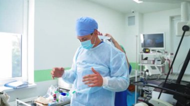 Modern ameliyathane. Asistanlar ameliyattan önce doktorun üniforma ve steril eldiven giymesine yardım ediyorlar. Sağlık hizmeti kavramı.