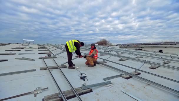 平らな屋根に太陽光発電所を設置する 労働者は美しい空の下で太陽光発電の太陽電池パネルのための金属の基礎を付けます — ストック動画