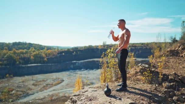 运动员运动后喝水的侧视图 年轻的运动员站在岩石山上 喝着瓶子里的水 — 图库视频影像