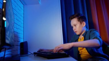 Oyun oynayan bir gencin portresi. Mutlu çocuk odasında oturup bilgisayarda çalarken klavye tuşlarına basıyor. Mavi akşam ışığı.