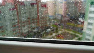 Yağmurlu havada cam pencere. Şehir arka planına yağmur damlaları düşüyor. Şehrin yağmurlu havasında daireden manzarayı izle.