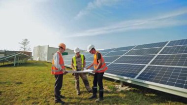 Güneş çiftliğinde proje planı olan erkek teknisyenler. Koruyucu üniformalı ve kasklı üç işçi parlak bir günde güneş panelleri kurulumu hakkında tartışıyorlar..