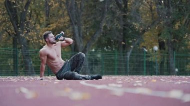 Üstsüz sporcu stadyumda su içiyor. Kaslı vücutlu sağlıklı bir sporcu dışarıda oturuyor ve antrenmandan sonra dinleniyor. Bir şişe suyu olan sportif bir adam..