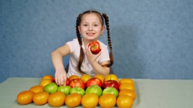 Küçük kız sulu elma yiyor. Masaya güzel yerleştirilmiş taze organik meyve. Mavi arka planda olgun meyveleri olan mutlu küçük kız..
